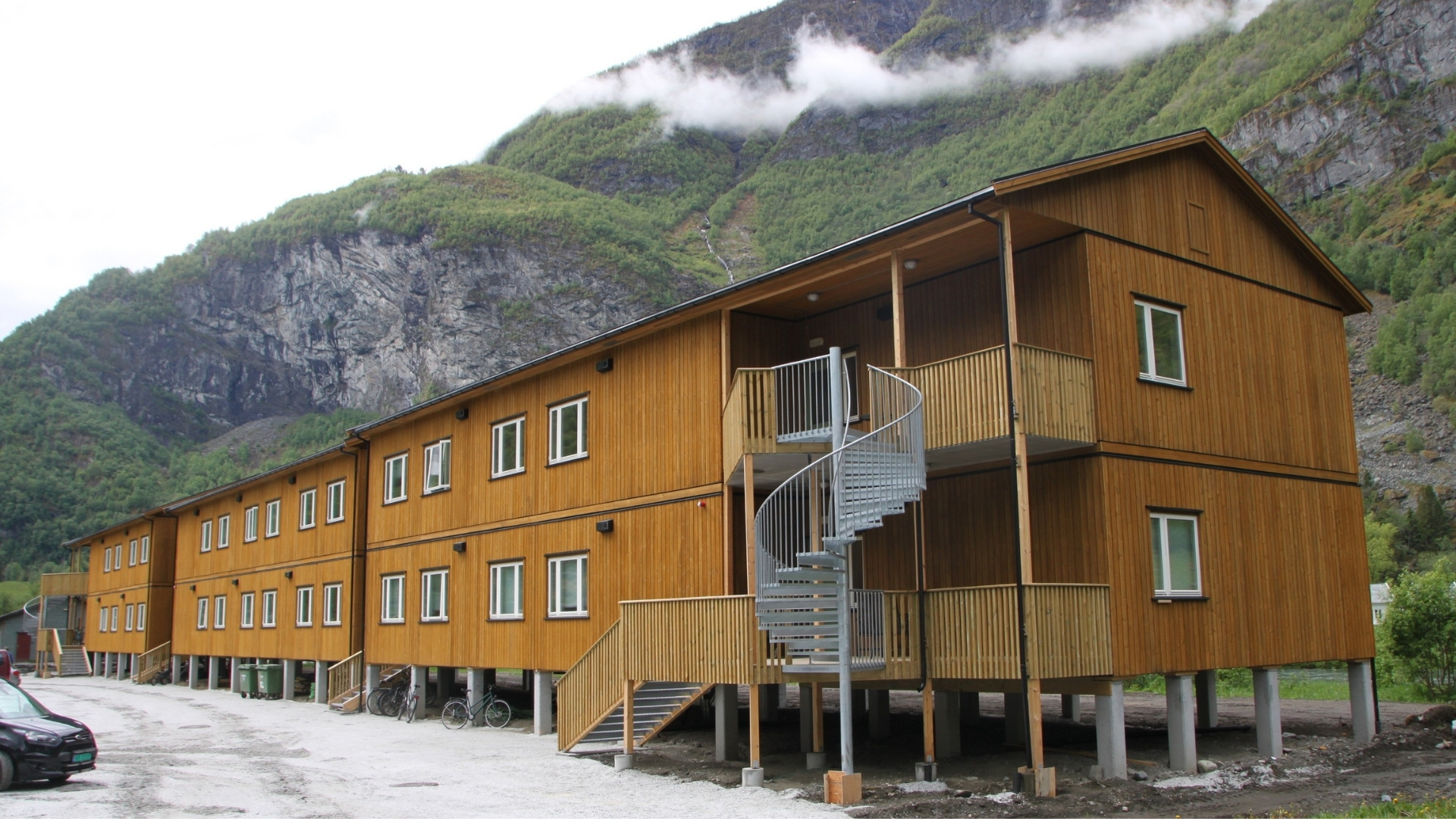 A long building with light brown wood cladding stands at the foot of a steep mountain.