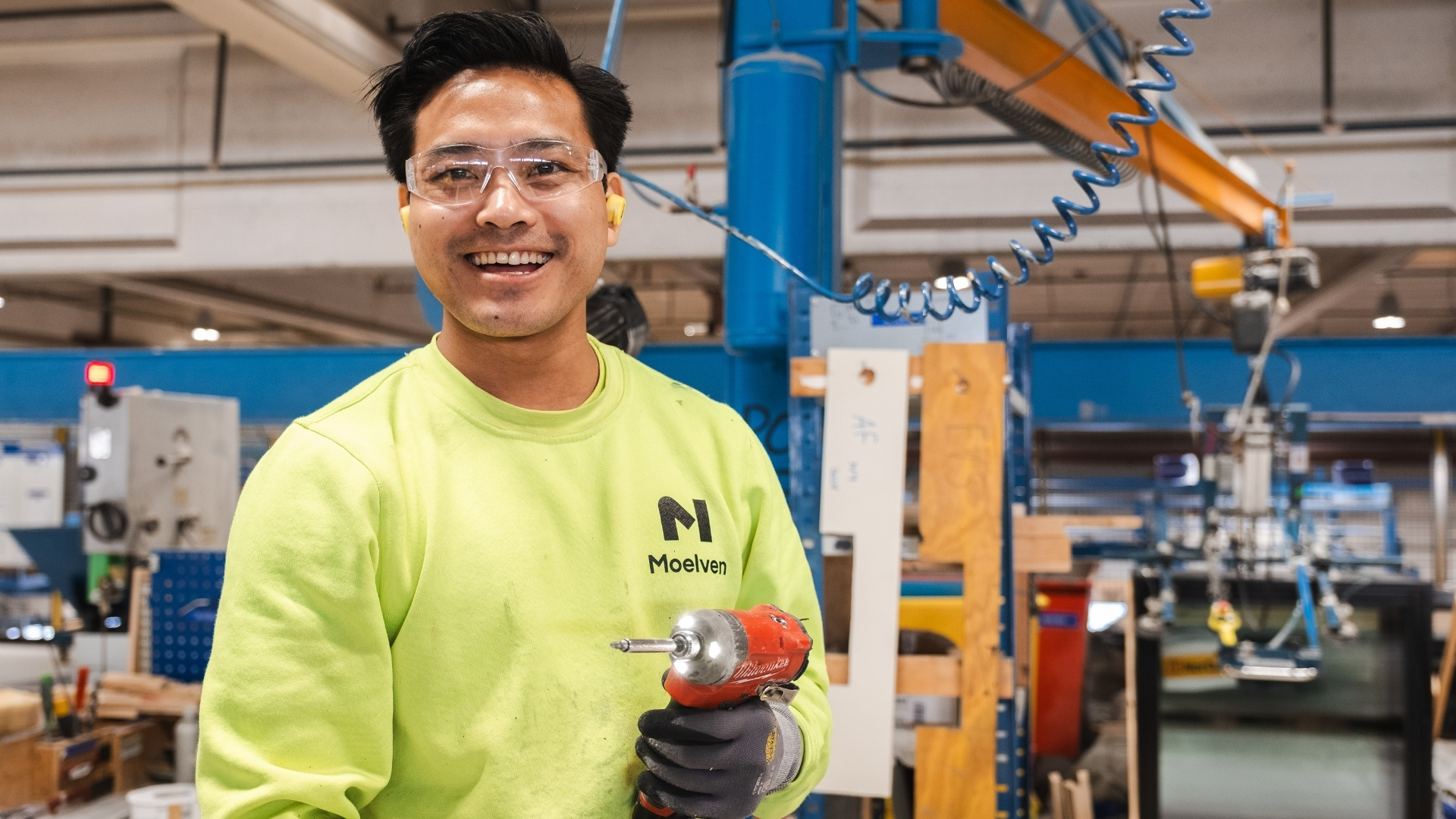 A smiling factory worker is holding a nail gun in their hand.
