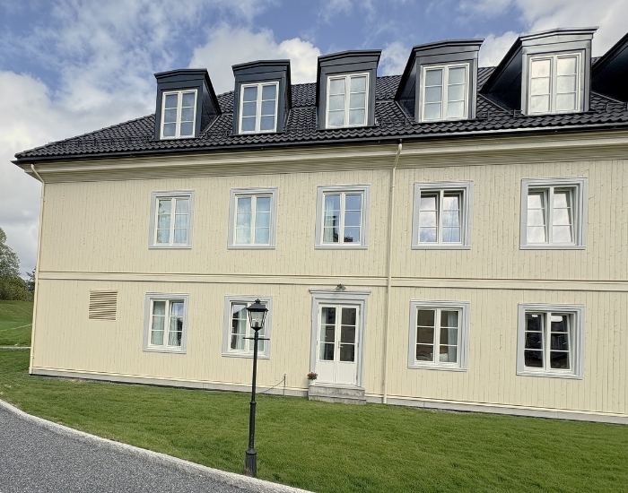 Lys gul trebygning med svarte arker på taket, omgitt av grønn plen og grussti i forgrunnen.