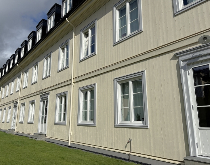 Lys gul bygning med hvite detaljer og symmetrisk vindusplassering, omgitt av grønn plen og trær, sett fra skrå vinkel